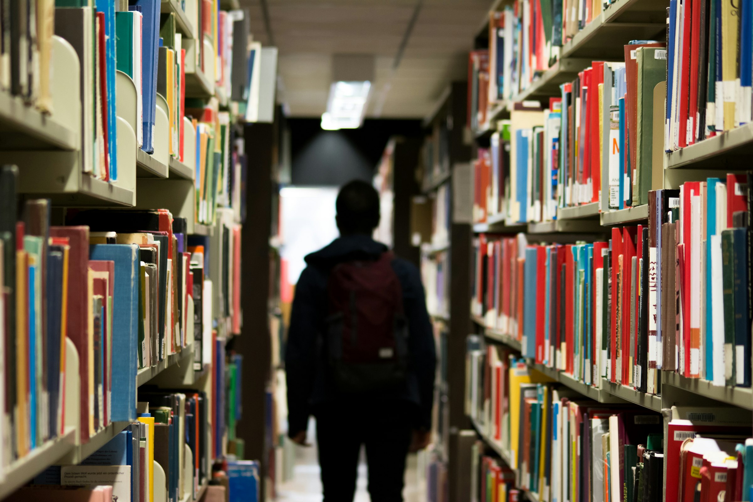 A student with a backpack walking down a library aisle between tall shelves of books